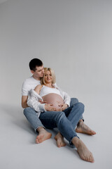 Family couple. A pregnant blonde Caucasian woman with her husband are sitting on a white cyclorama and hugging, gently touching their stomachs. They wear jeans and a white T-shirt.