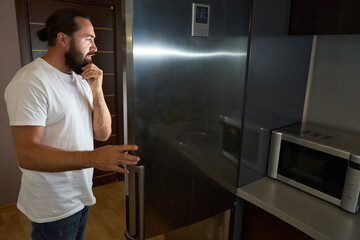 Middle-aged male stands, thinking, in front of a refrigerator