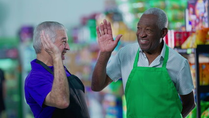 Happy diverse senior employees celebrating success with high-five standing in supermarket aisle. Caucasian manager engaging with workforce teamwork with African American colleague