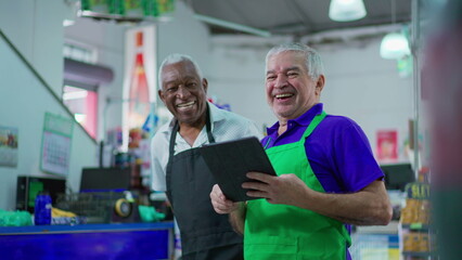 Obraz premium Joyful diverse Brazilian senior staff workers of supermarket chain smiling at camera with table and uniforms. African American older employee and a caucasian person laughing together
