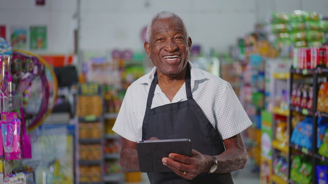 Joyful Happy Brazilian senior employee of supermarkert wearing apron and holding tablet device, portrait of a black older male staff depicting job occupation at retail store