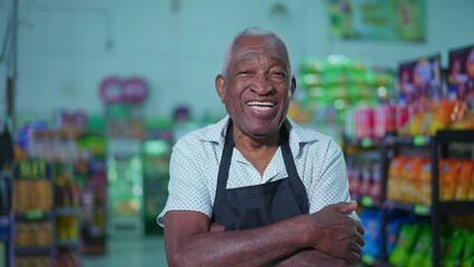 Portrait of a joyful African American senior employee of supermarket wearing apron and smiling at camera inside grocery store aisle and arms crossed