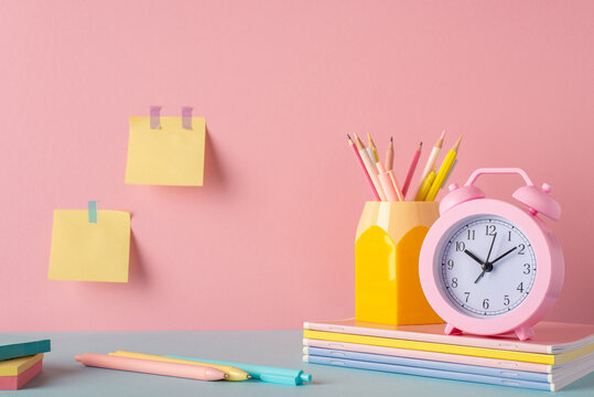 Back to school concept. Photo of school supplies on blue table pencil holder alarm clock over stack of notebooks pens and sticky note paper attached to pink wall