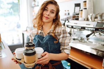 Portrait of a young woman barista holding takeaway coffee in her hands. Owner of the coffee shop stands behind the bar in an apron and gives coffee to go. Takeaway food. Business concept.