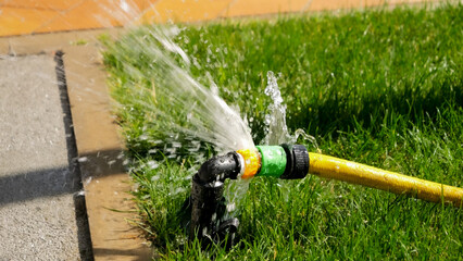 Closeup of water leaking and flowing on green grass lawn through damaged hose pipe. Water waste, garden equipment, gardening.