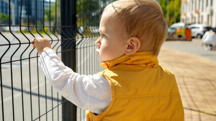 Portrait of sad baby boy standing alone at the fence on backyard playground. Upset children,...