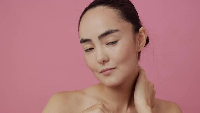 Young beautiful asian woman with clean skin looking at the camera and gently touching her neck with both hands on a pink background. Concept of skin care for health and beauty