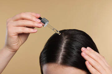 Woman applying essential oil onto hair roots on beige background, closeup
