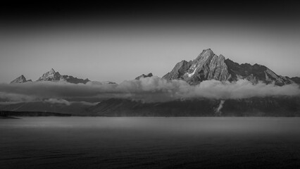 Black and White Photo of Clouds hanging over Jackson Lake as the sun rises and lights up the tallest peaks of Grand Teton National Park, Wyoming, USA