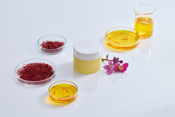 On the white table, unbranded jar decorated with flowers and few petri dishes containing yellow fluid and saffron. Gentle skin care concept