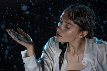 young woman in the rain, getting wet through, wet clothes and water running down her face. beautiful brunette catches raindrops in her palm. portrait in aqua studio