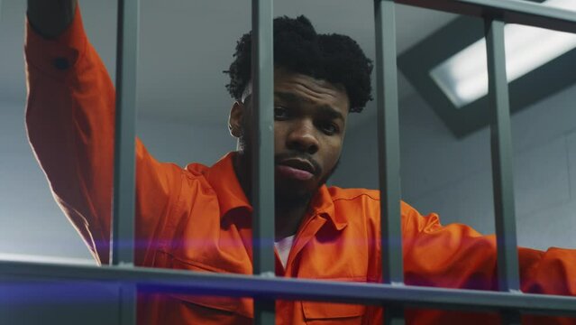 African American man in orange uniform leans hands on prison cell bars and looks at camera. Depressed prisoner serves imprisonment term in jail. Murderer in correctional facility or detention center.