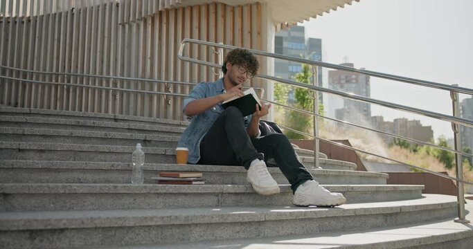Young indian guy student making notes in a notebook while sitting on a stairs outdoor