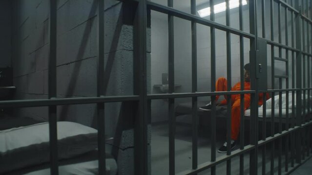 Two neighbor prisoners serve imprisonment term in prison cells. African American criminal in orange uniform sits on jail bed and looks at barred window. Gloomy inmates in detention center. Dolly shot.