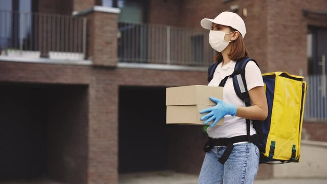 Side View of the Delivery Woman Holding Paper Box, Wearing Protective Mask and Gloves, Carrying the Yellow Backpack. Delivery While Pandemic. Woman Worker Delivery While Covid. Postal, Postwoman