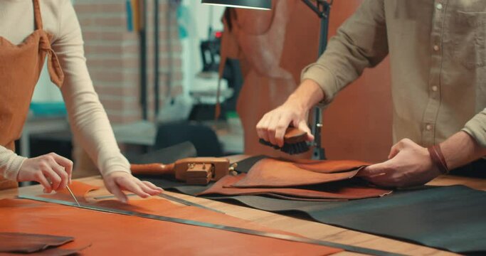 handsome man and red-haired woman working together in a manufacture workshop female uses awl marks a piece of leather, man cleaning brishing material Slow motion Family business