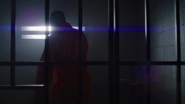 Depressed African American man in orange uniform stands from jail bed, walks at prison cell and looks at barred window. Sad prisoner serves imprisonment term. Murderer in gloomy correctional facility.