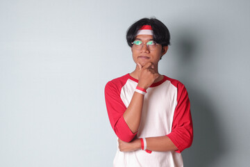 Portrait of attractive Asian man in t-shirt with red and white ribbon on head, standing against gray background, thinking about question with hand on chin