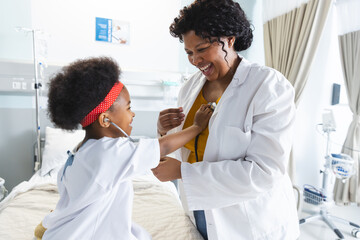 African american female doctor playing with girl patient using stethoscope at hospital - Powered by Adobe