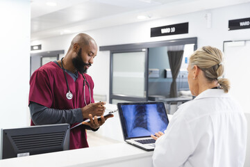 Diverse male and female doctors discussing work and using laptop in reception at hospital