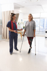 Caucasian female physiotherapist and senior woman with artificial leg walking with crutches