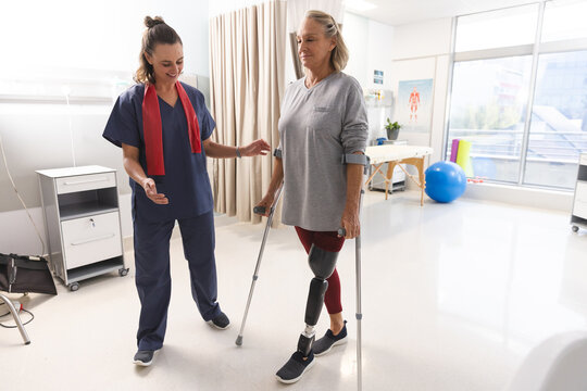 Caucasian female physiotherapist and senior woman with artificial leg walking with crutches