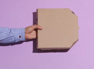 Man's hand in shirt holding craft cardboard pizza box on purple pastel background with shadow.