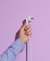 Man's hand in shirt holding electric plug on purple pastel background with shadow.