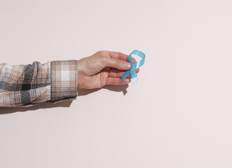 Male hand in shirt holding blue prostate cancer awareness ribbon on beige pastel background