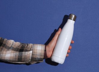Man's hand in shirt holding white thermos bottle on blue background with shadow