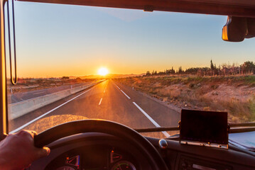 View from inside the cab of a truck driving on a highway with the early morning sun behind some mountains.