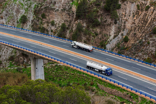Two fuel tanker trucks crossing on a highway over a bridge, aerial view.