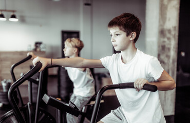 Fototapeta na wymiar Two teenager boys are doing exercise with airbike in the gym. Cardio, strength training, fitness concept