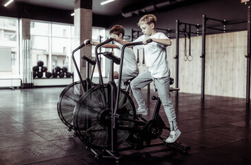 Fototapeta na wymiar Two teenager boys are doing exercise with airbike in the gym. Cardio, strength training, fitness concept