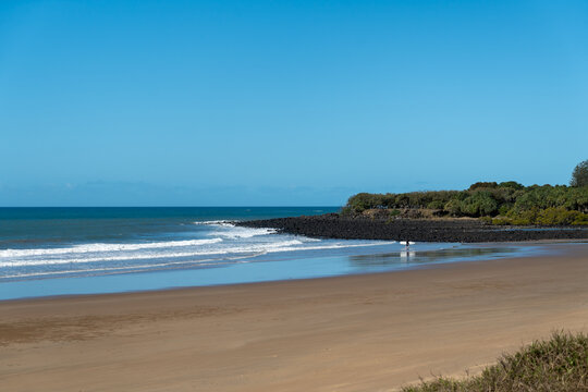 Surf beach with sand, rock wall, headland with trees and view to the horizon. Bargara, Queensland, Australia.