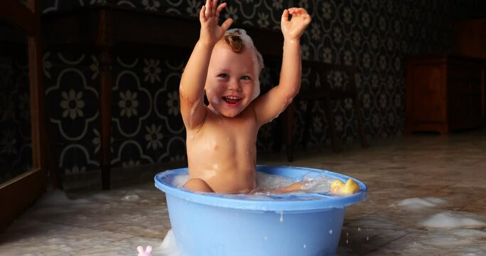 Cheerful and mischievous little boy in baby bathtub creates playful splashes of water with his enthusiastic slaps. Slow motion footage captures his vibrant emotions and carefree enjoyment