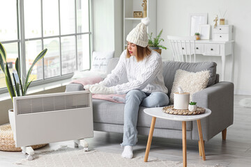 Frozen young woman in winter clothes warming near radiator at home
