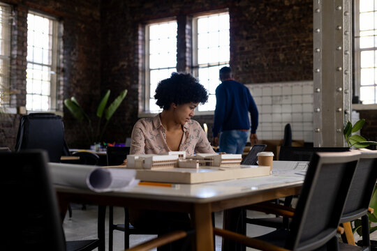 Biracial female architect sitting at desk using tablet and looking at building model in office
