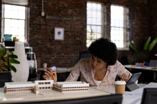 Biracial female architect sitting at desk using tablet and looking at building model in office
