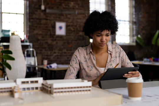 Biracial female architect using tablet sitting at desk with building model in office