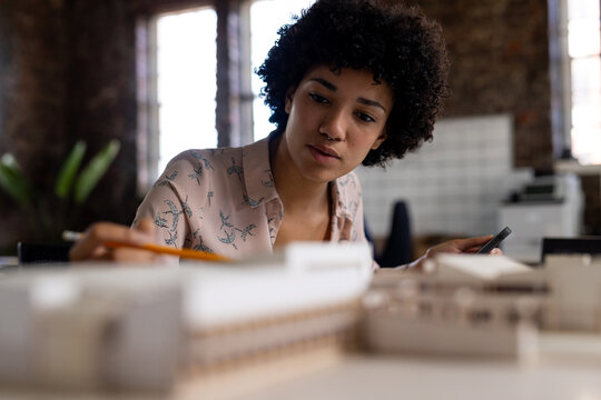 Biracial female architect sitting at desk using smartphone and looking at building model in office