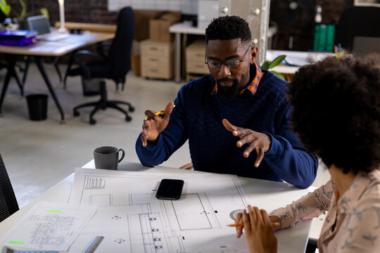 Focused diverse male and female architects discussing blueprints sitting at desk in office