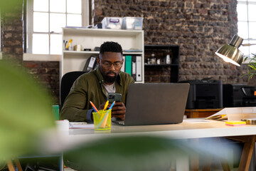 African american casual businessman sitting at desk using laptop and smartphone at office