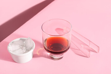Glass of rum, bowl with ice cubes and tongs on pink background