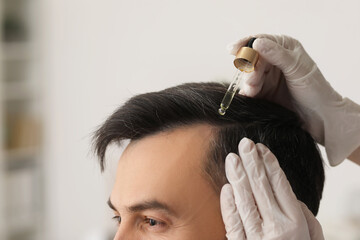 Doctor applying serum on young man's hair in clinic, closeup
