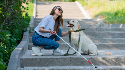 Blind young blonde petting a guide dog on a walk in the park. Woman with tactile cane at the...
