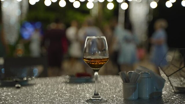 Unfinished glass of alcohol and ceramic menage stands on festive shiny table at party. Blurred people in summer dresses are dancing under garlands with silver tinseland during disco outdoor.