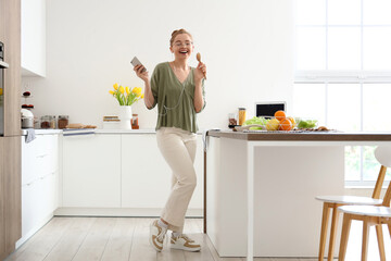 Pretty young woman listening to music, dancing and using wooden spoon as microphone in light kitchen