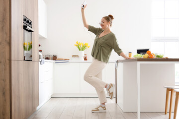 Happy young woman listening to music and dancing in light kitchen