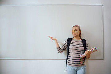 Happy female student pointing at blank whiteboard in classroom and looking at camera.